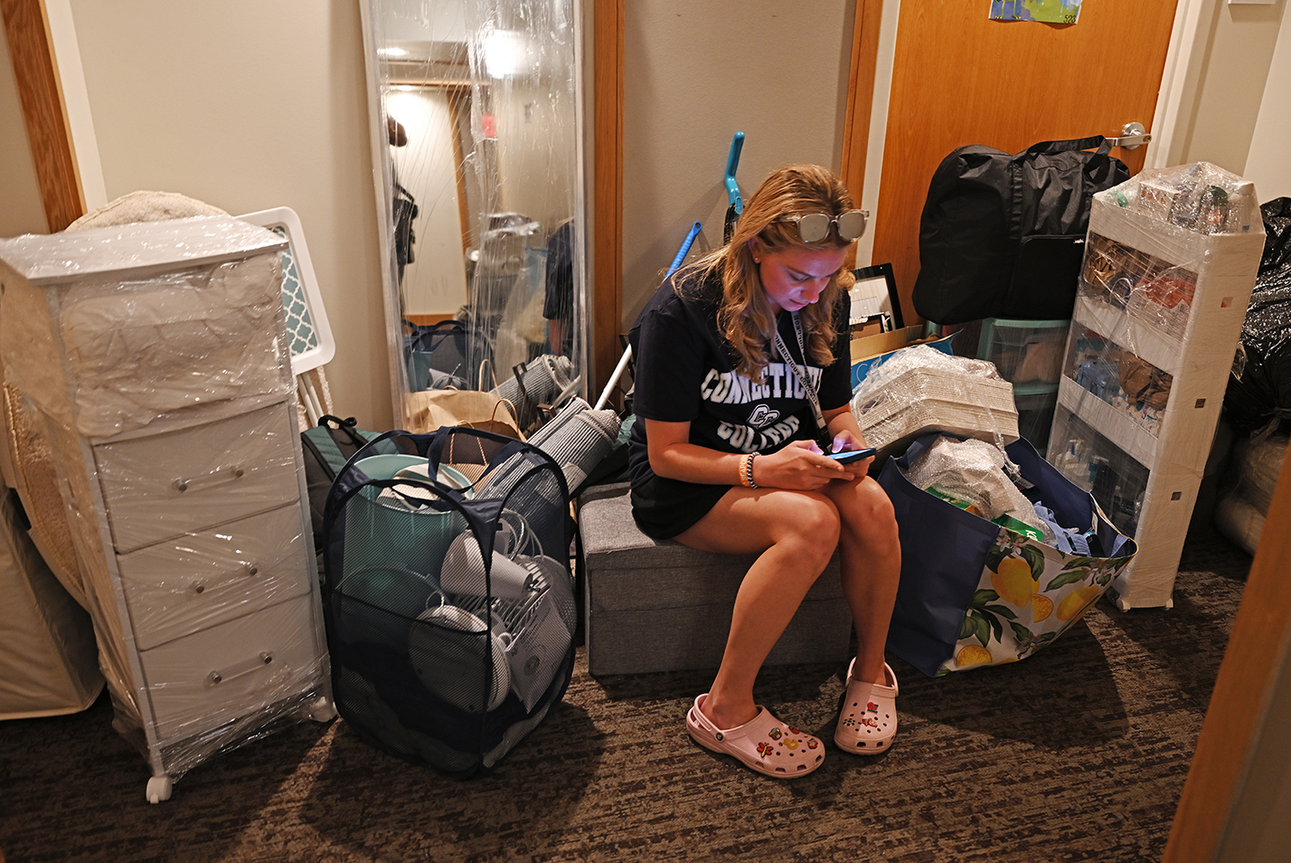 A student sits on a large pile of belongings waiting to move into a dorm room.