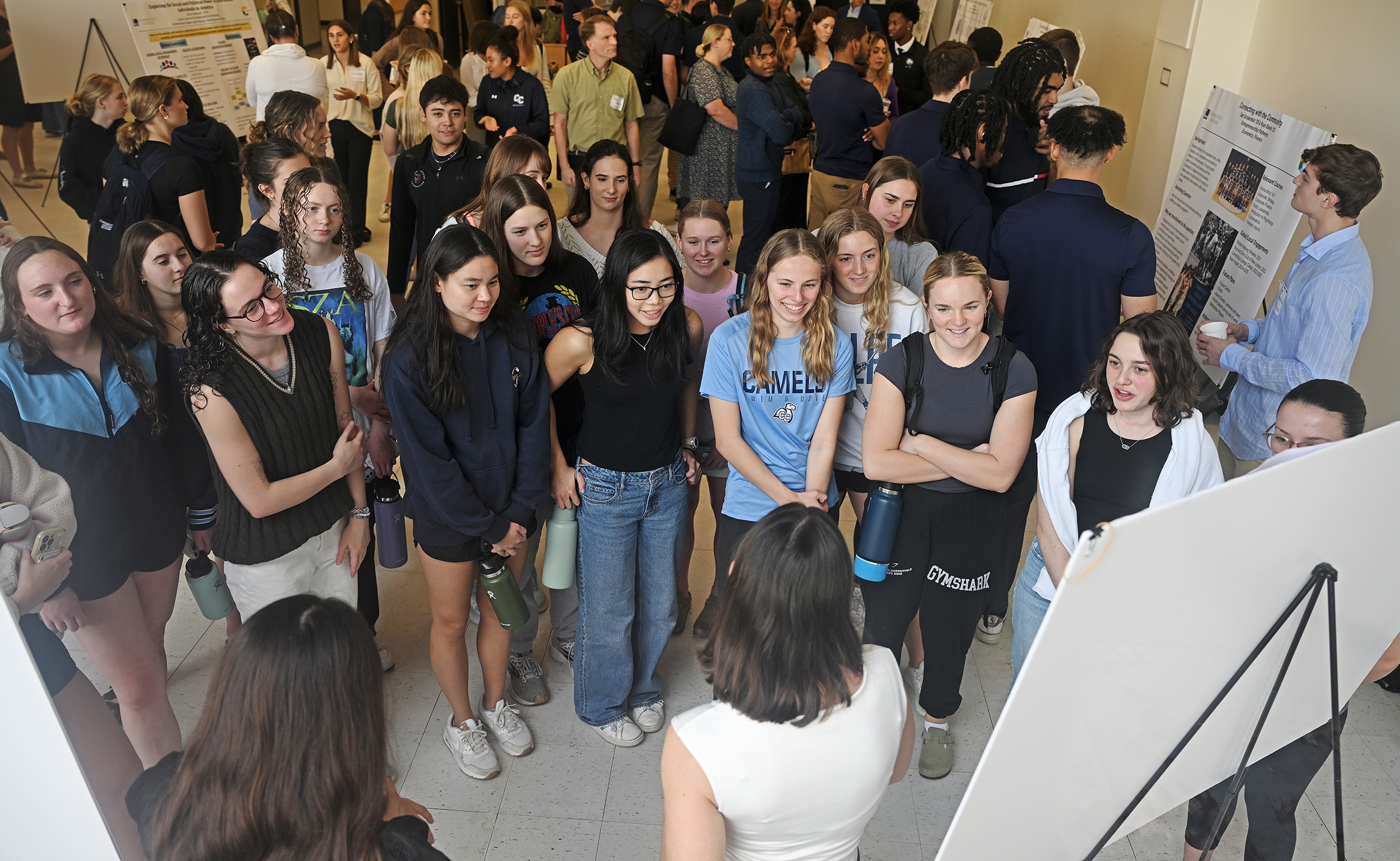 Students crowd around another presenting research results on a large poster board.